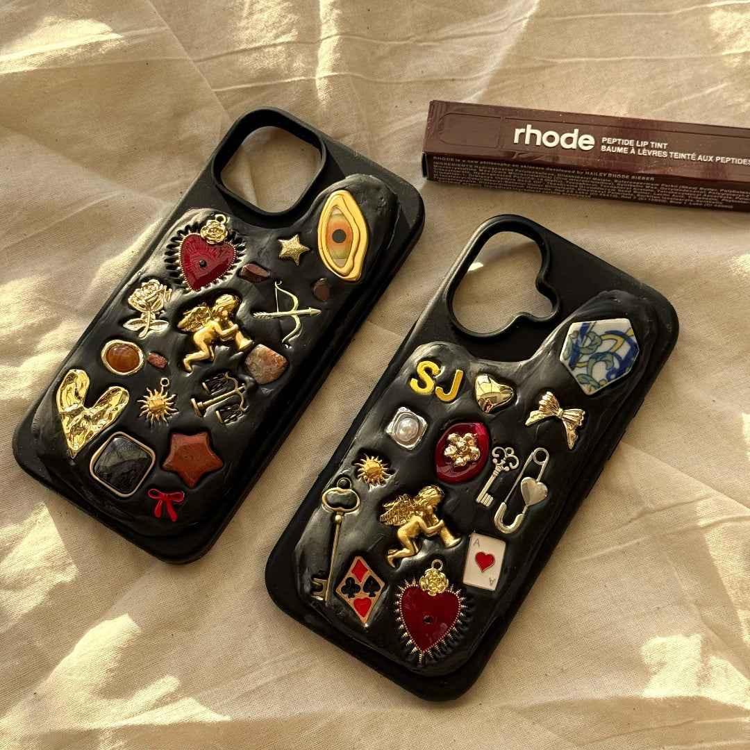 Two black phone cases with decorative pins on a beige surface, with 'rhode' product in the background.