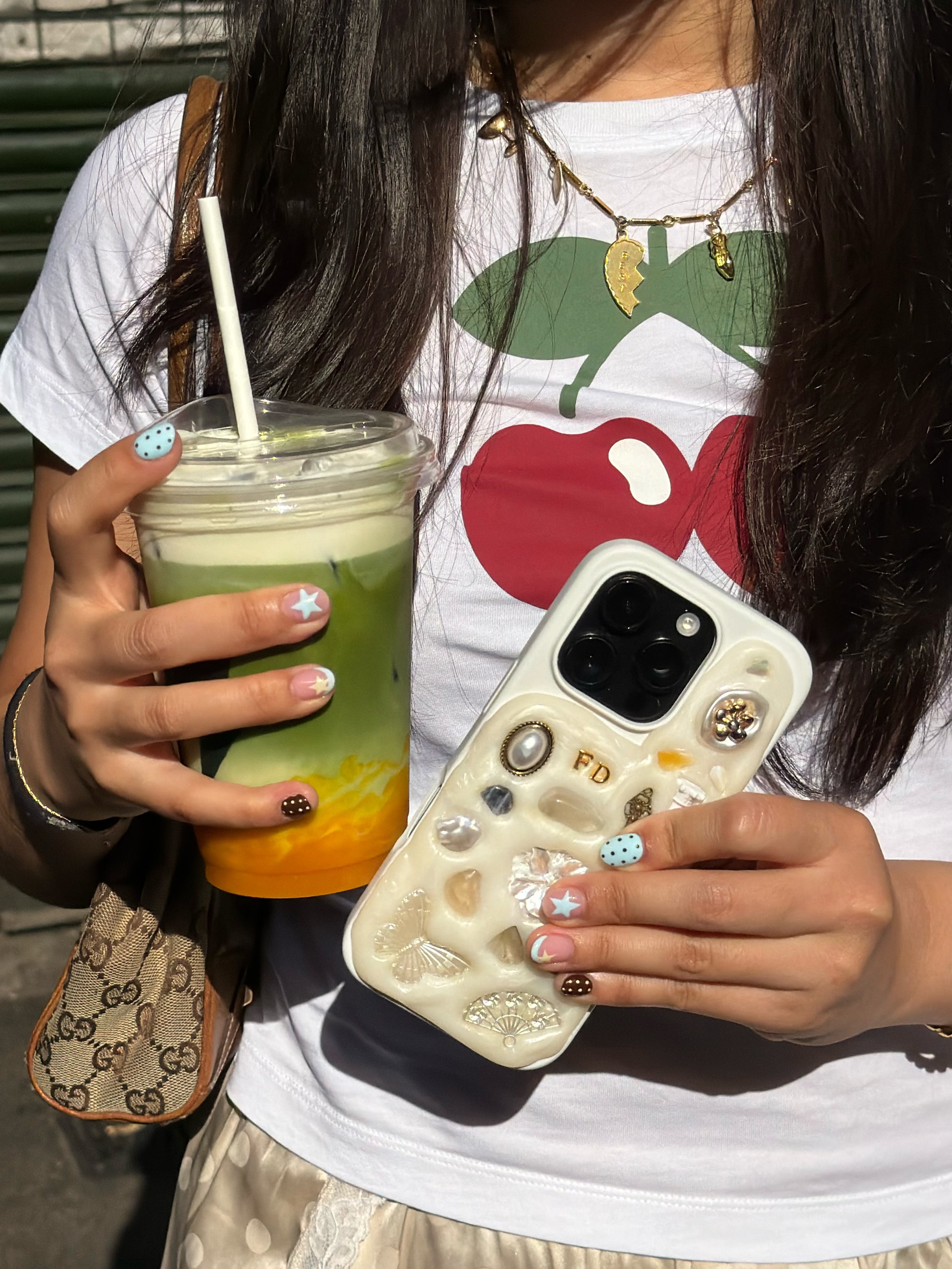 Person holding a drink and phone with decorative case, wearing a white shirt with a red and green design.
