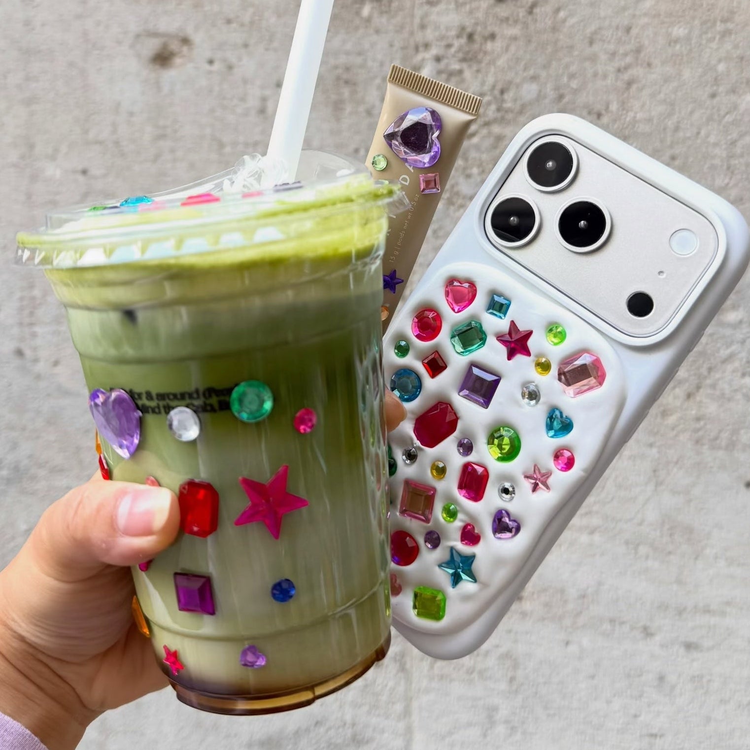A hand holds a decorated iced matcha drink in a clear plastic cup and a white smartphone case covered with colorful rhinestones, against a textured gray wall.