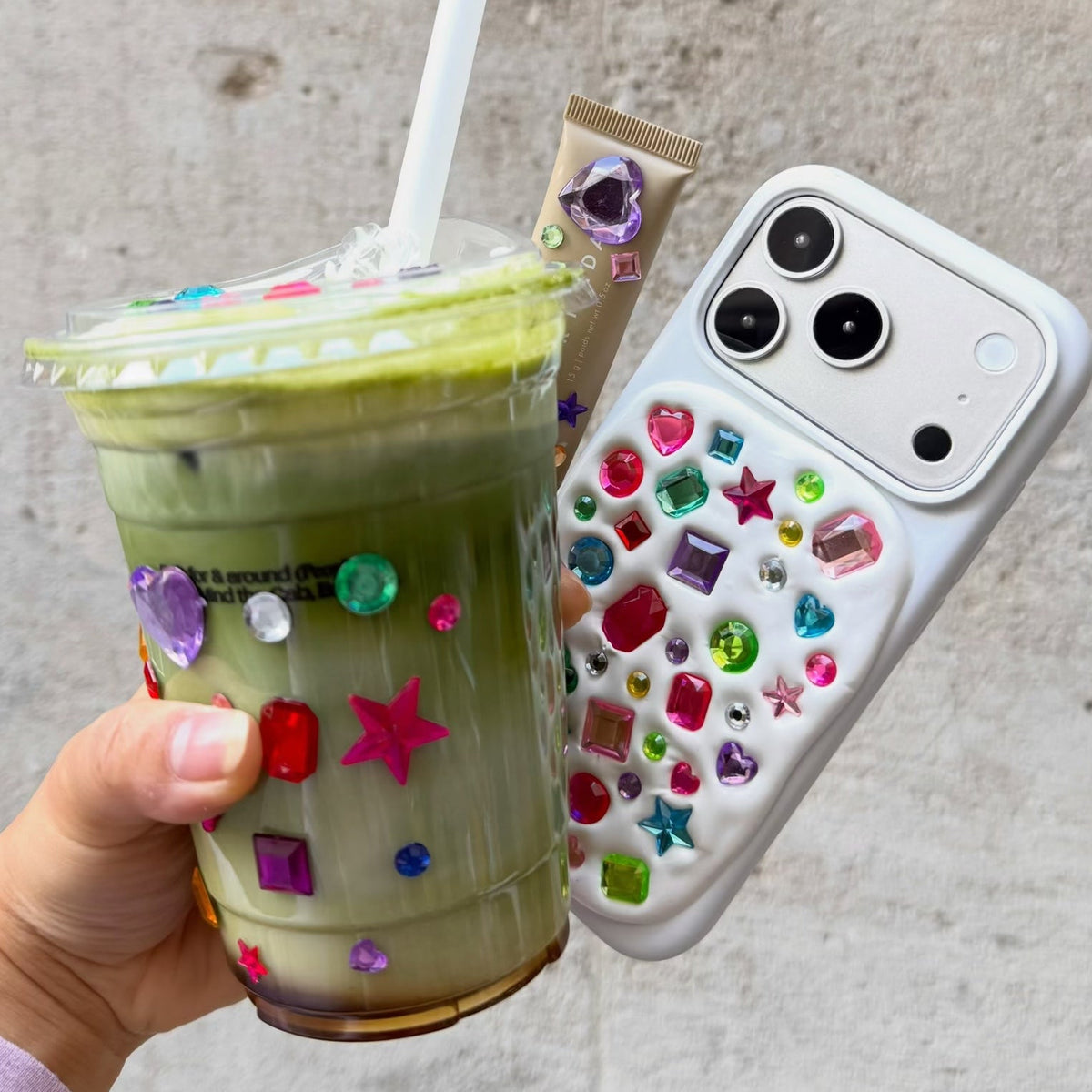 A hand holds a decorated iced matcha drink in a clear plastic cup and a white smartphone case covered with colorful rhinestones, against a textured gray wall.