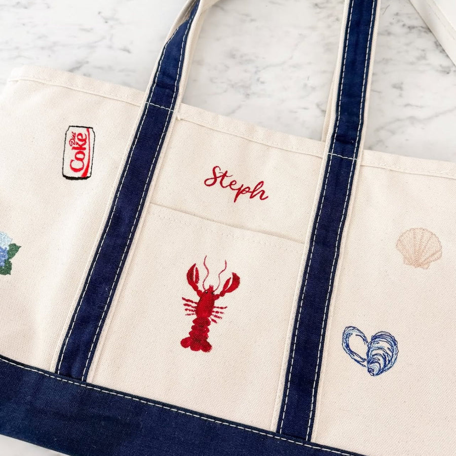 A cream canvas tote bag with navy blue trim and handles, featuring embroidered nautical-themed designs including a red lobster, blue seahorse, beige seashell, and the name 'Sophia' in red script.