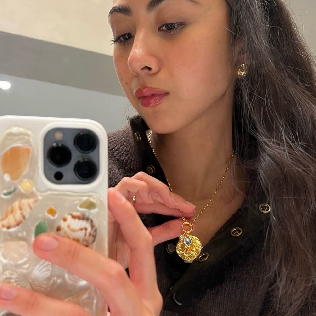 A woman taking a mirror selfie while wearing a gold pendant necklace with a detailed shell design and a blue stone accent. She is holding a white phone case decorated with seashells.