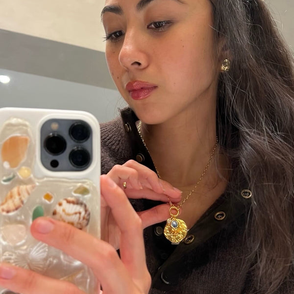 A woman taking a mirror selfie while wearing a gold pendant necklace with a detailed shell design and a blue stone accent. She is holding a white phone case decorated with seashells.